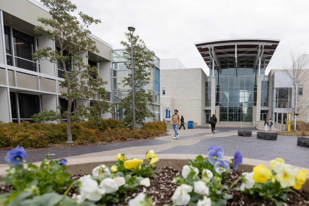 A view of Stockton University's Campus Center in early Spring. Students walk in the background with flowers blooming in a large planter in the foreground.