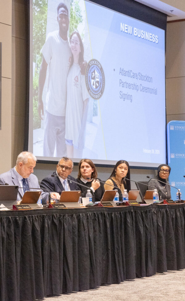 Several members of the Stockton Board of Trustees sit at a table presenting Stockton's partnership with Atlanticare which is featured on a projection screen behind them.