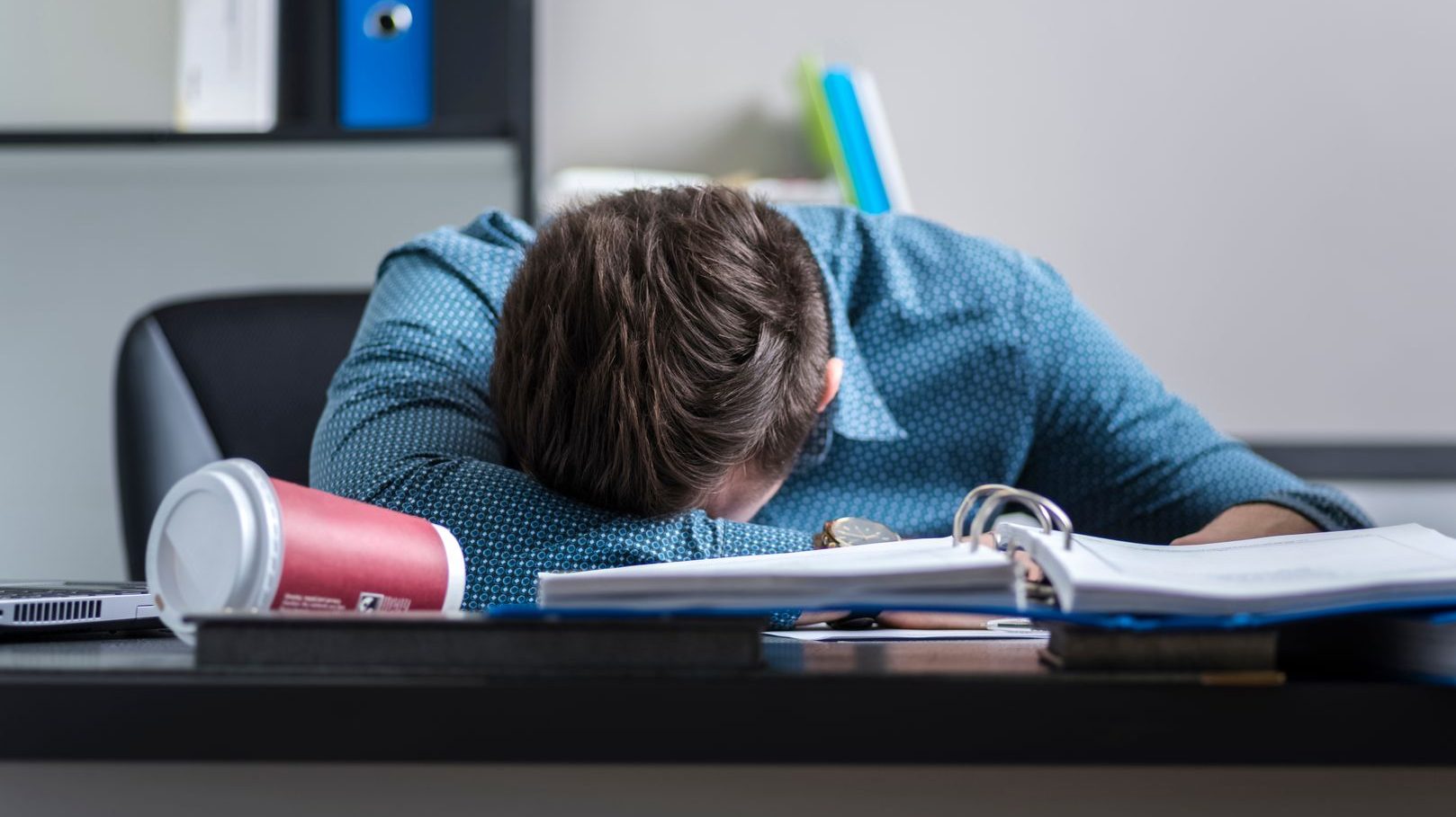 Tired male worker asleep at a desk. A cup of coffee is knocked over next to him.