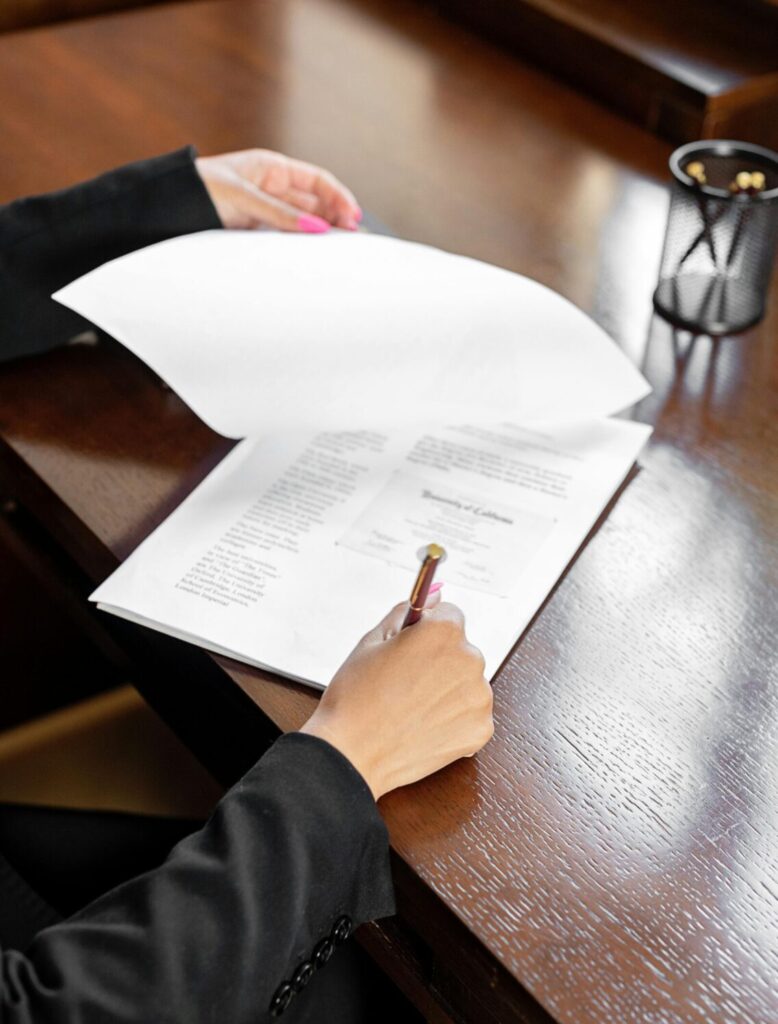 A person, out-of-frame, sits at a desk, their hands can be seen holding and signing a paper.
